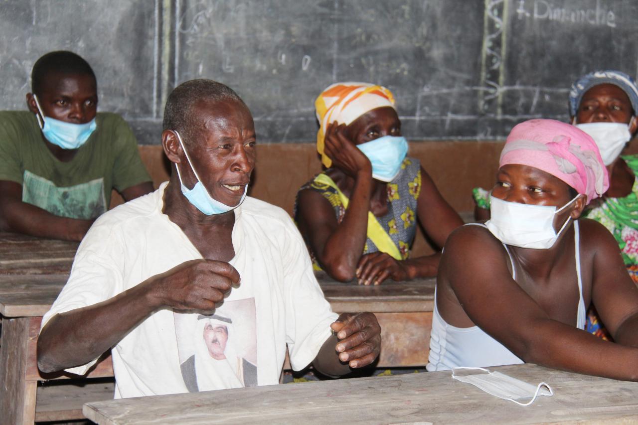 An older man is sitting at a wooden table in a classroom and speaking with a face mask pulled down under his chin. Several people, all wearing face masks, are sitting next to him and behind him.