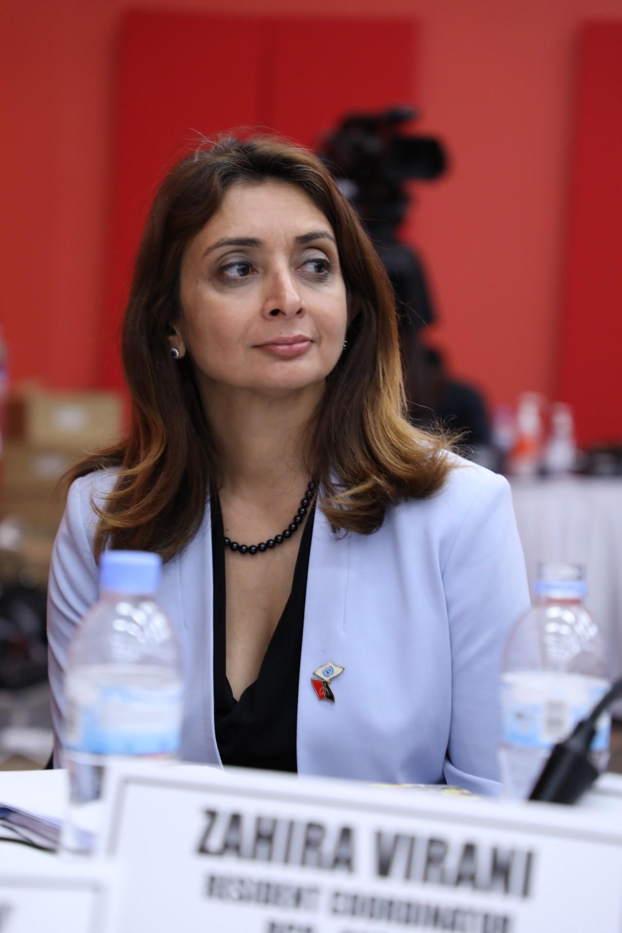 A woman in a light purple blazer sits in front of a red-colored wall. 