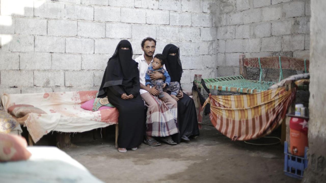 Un homme tenant un jeune enfant sur les genoux est assis sur une banquette, entourée de deux femmes portant le voile intégral, dans une pièce meublée de façon rudimentaire et dont les murs en brique sont grossièrement peints en beige.