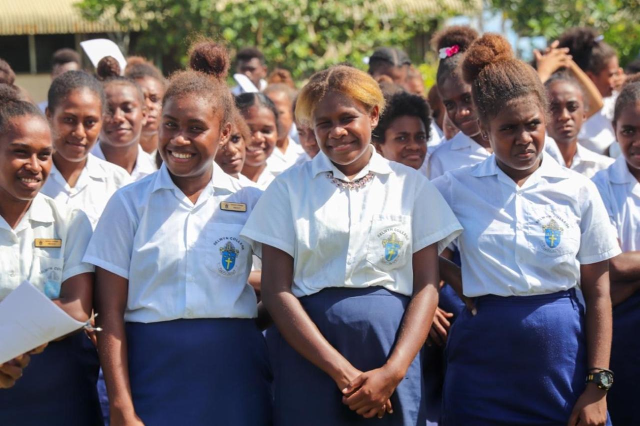 Teen girls in school uniforms - blue skirts and white-button downs - fold their hands and smile.