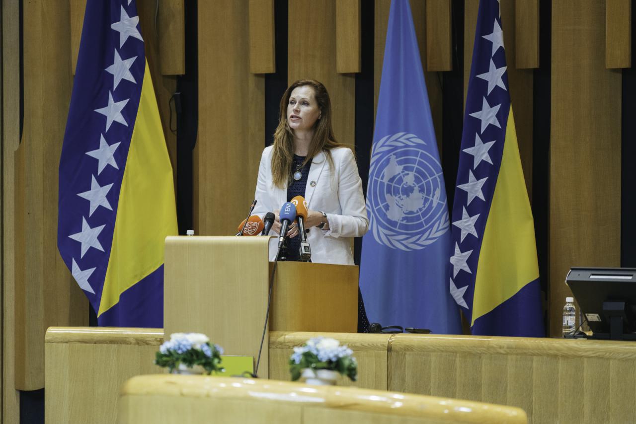 A woman in beige suit is speaking into a microphone at a podium.
