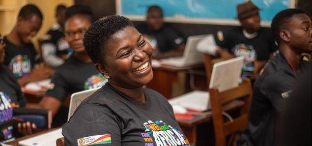 Улыбающаяся молодая женщина с короткими волосами в футболке с логотипом Africa Code Week сфотографирована в классной комнате.