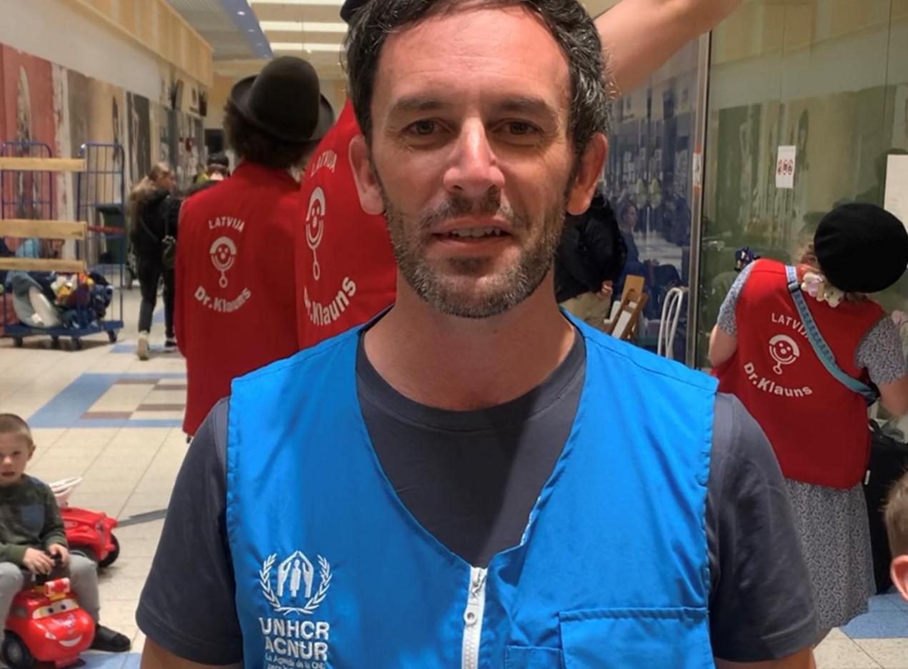 In an indoor space, a man in a blue vest looks at the camera with determination and a group of people in red vests and a group of refugees stand in the background.