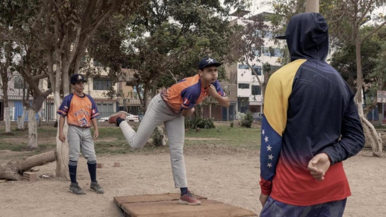 A young boy in an orange uniform pitches a baseball while two other players