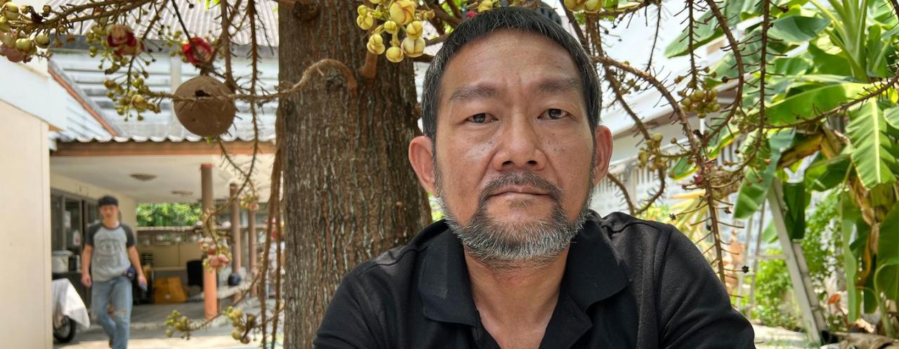 A man in a black shirt and a goatee sits in a place while bamboo poles are in the background