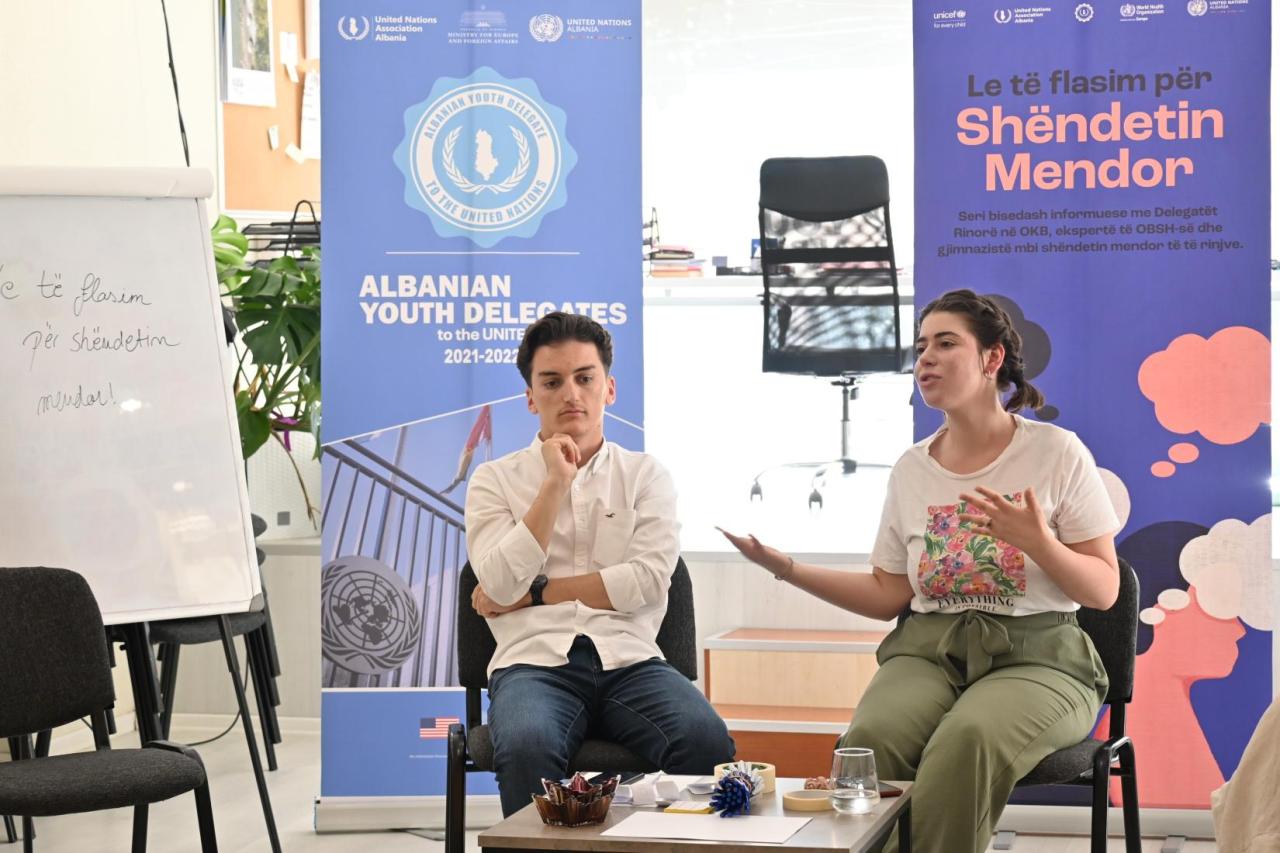 A young woman in a white shirt and green pants gestures as she talks while another young man seated next to her listens closely