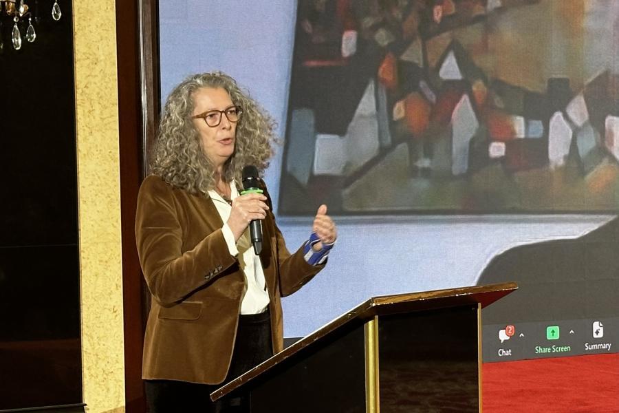 A woman in a brown coat with curly grey hair and glasses speaks into a microphone at a podium