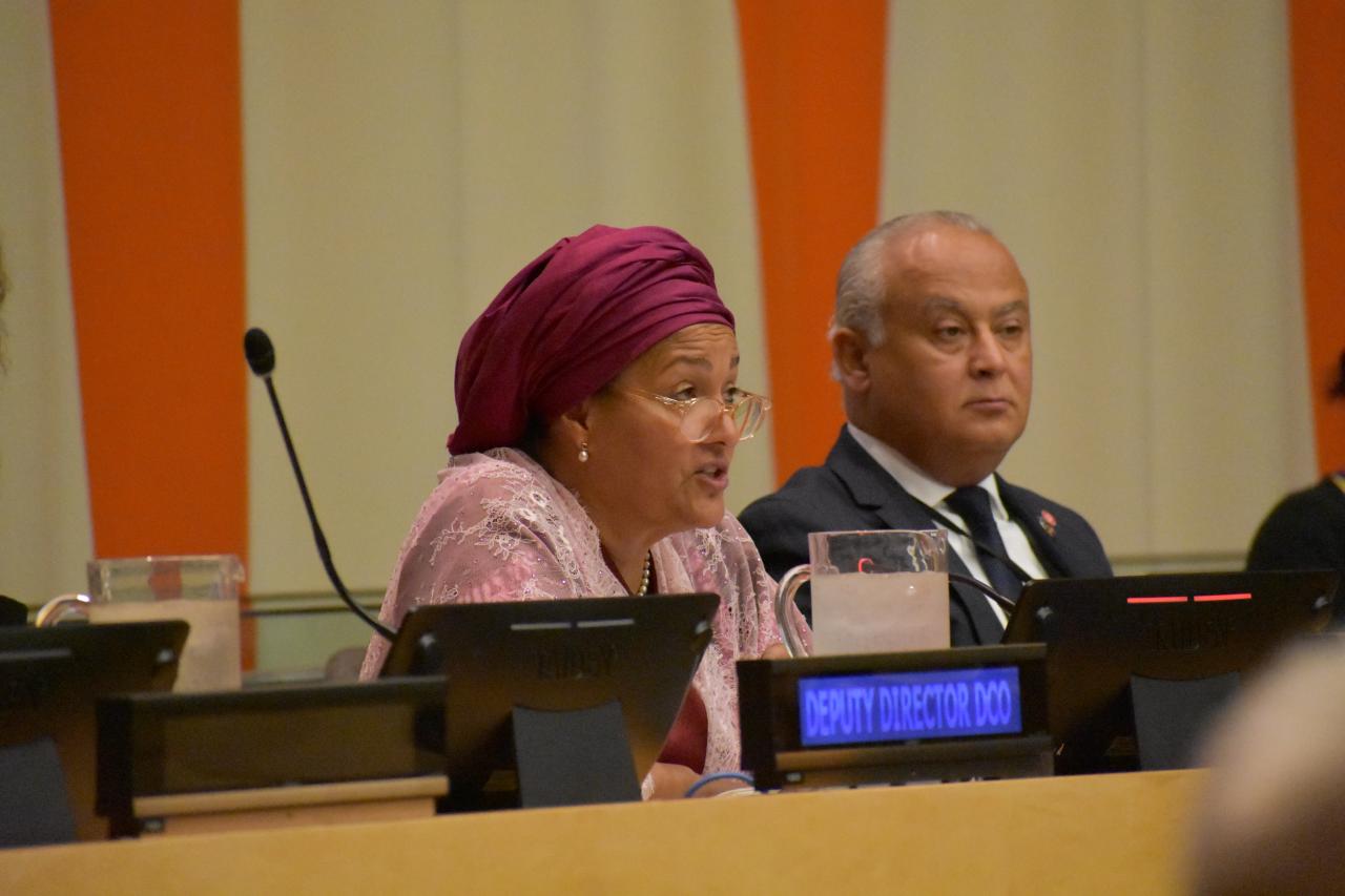 A woman in a red headscarf and pink shirt speaks into a microphone as a man in a black suit listens on
