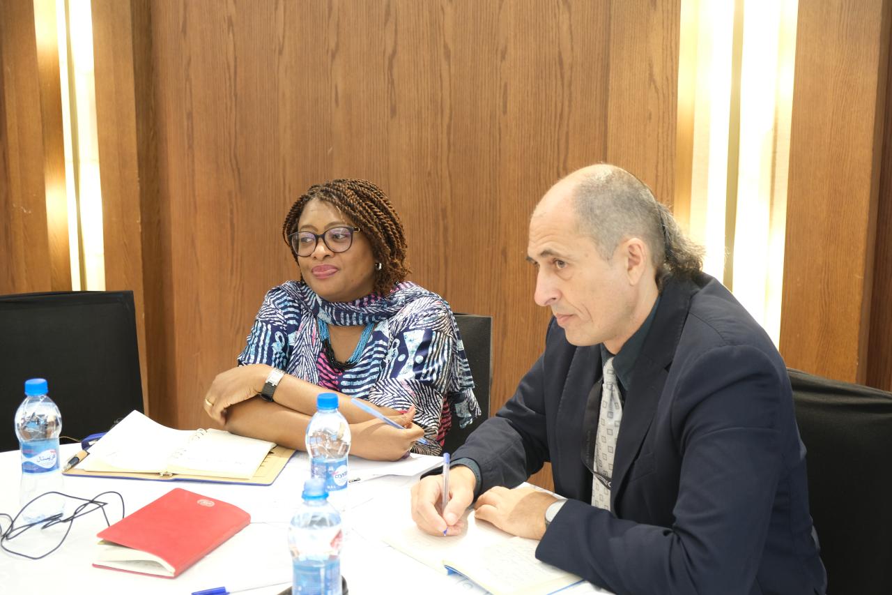 A man in a black suit, seated next t a woman in a blue dress listens to discussions.