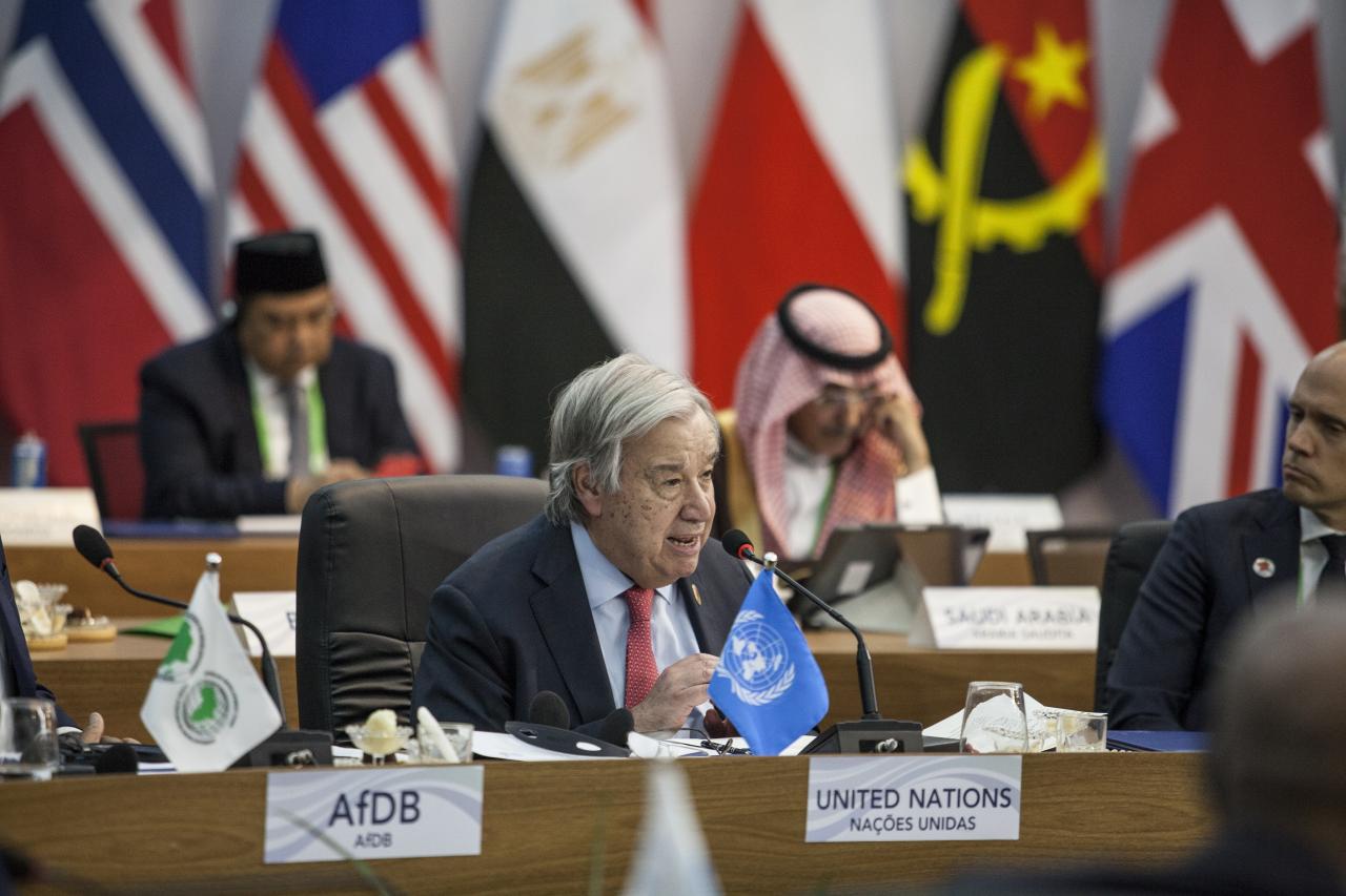 UN Secretary-General Antonio Guterres in a blue suit and red tie speaks into a microphone