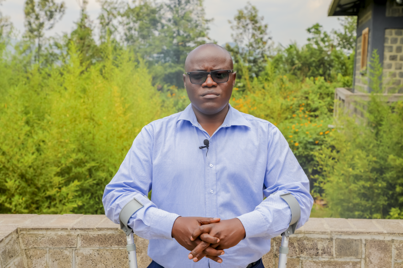 A man in a blue shirt wearing dark glasses stands on a pair of crutches against a lush green background