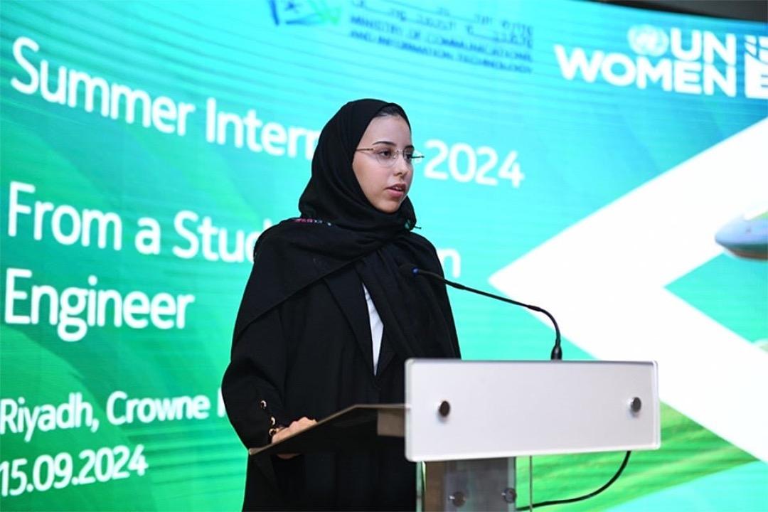 A woman in a black full-covered attire and spectacle speaks into a microphone against a colourful LED screen