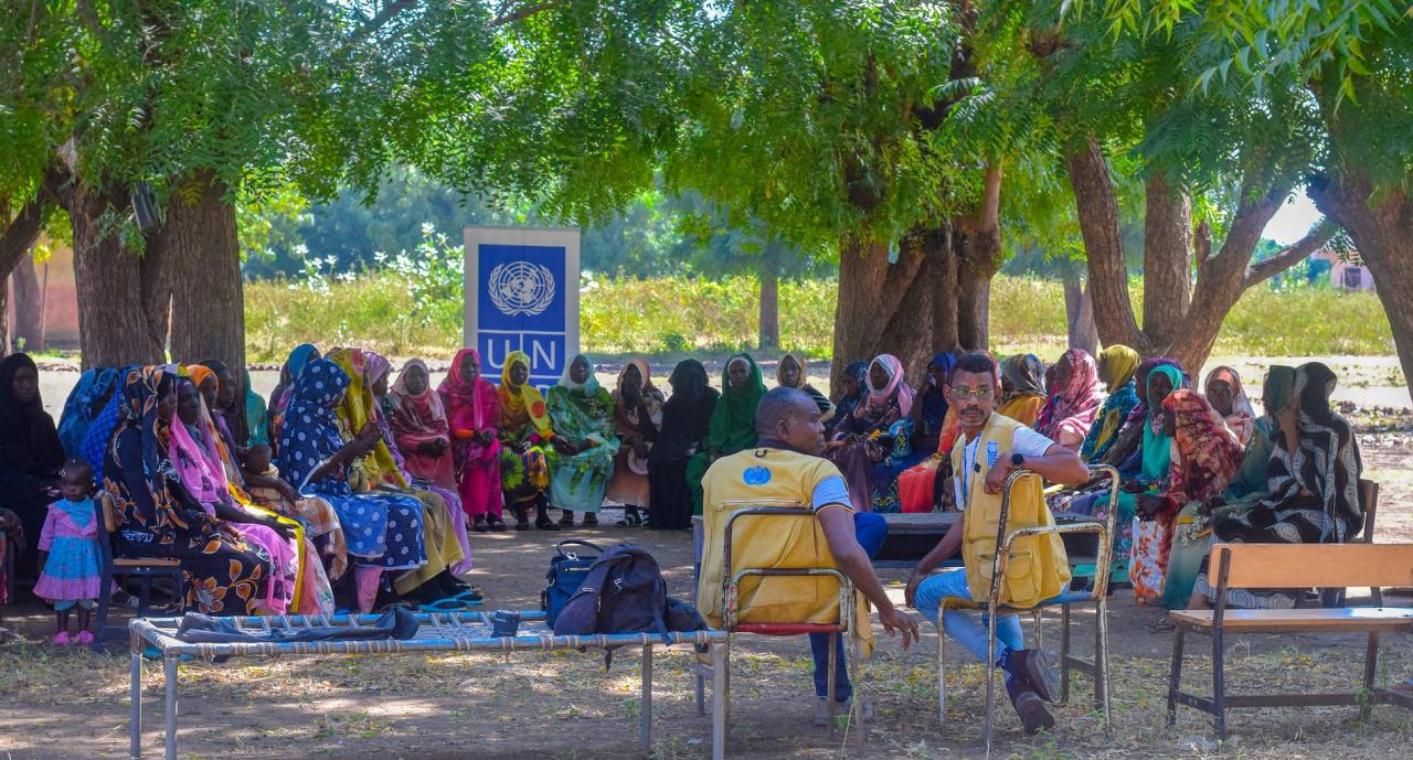 A group of about twenty or so women gather in a common place among trees. Two men in yellow vests with the UNDP logo sit across from them.