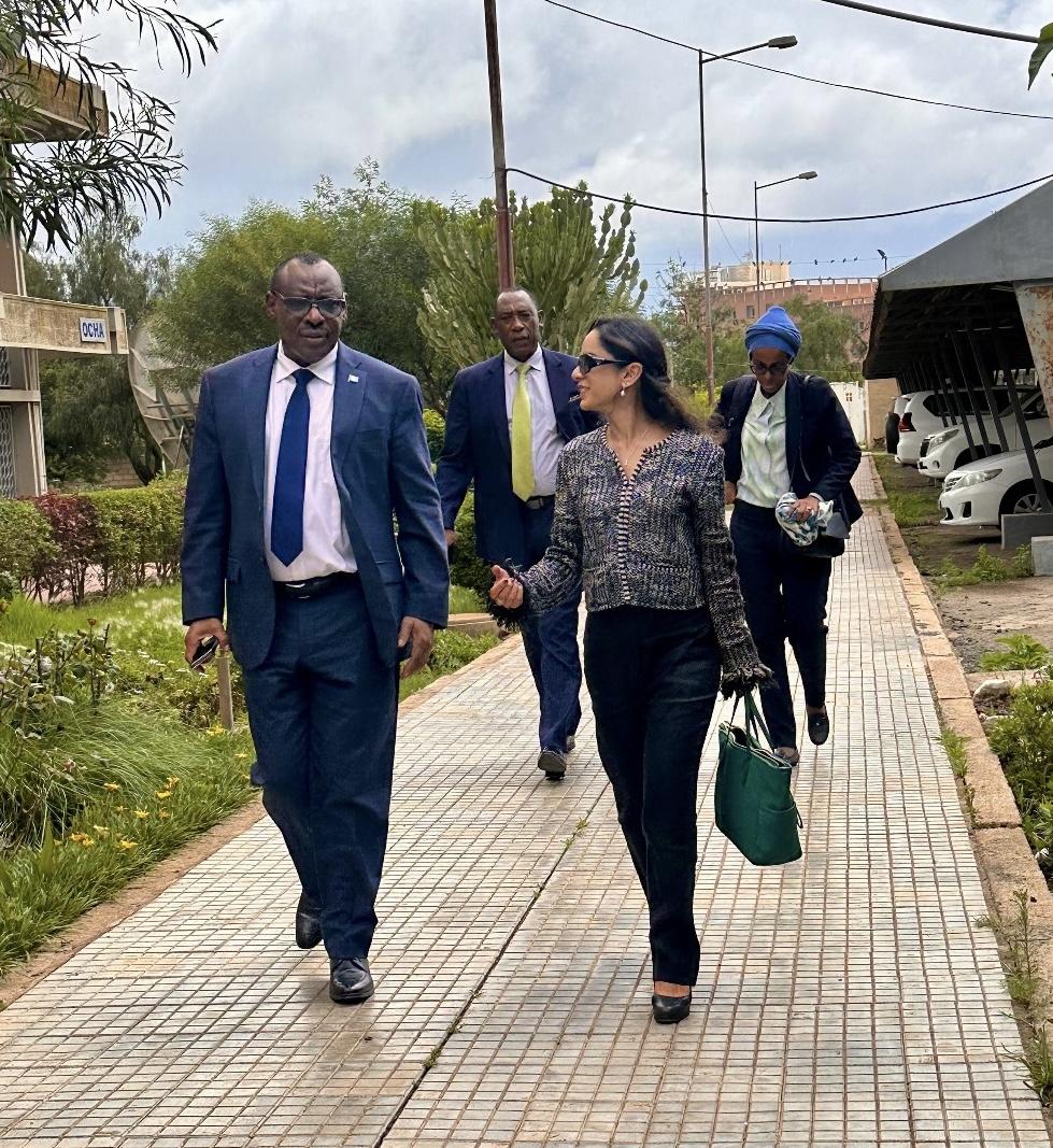 A woman in a black formal dress and sunglasses walks and talks to a man in a navy suit. They are followed by two men in suits.