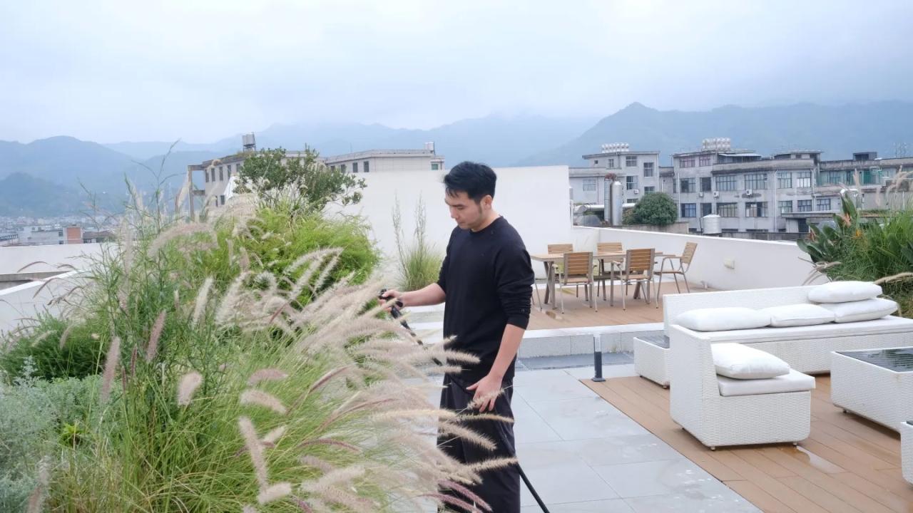 Una persona riega y cuida plantas en una terraza ajardinada en la azotea, con mobiliario exterior y vistas urbanas y montañosas al fondo.