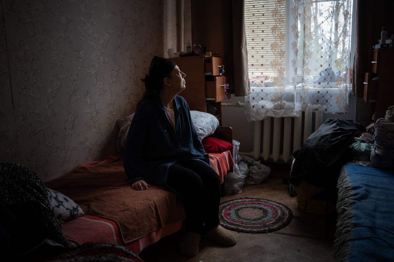 A woman in Ukraine sits in a bed and looks out a window.