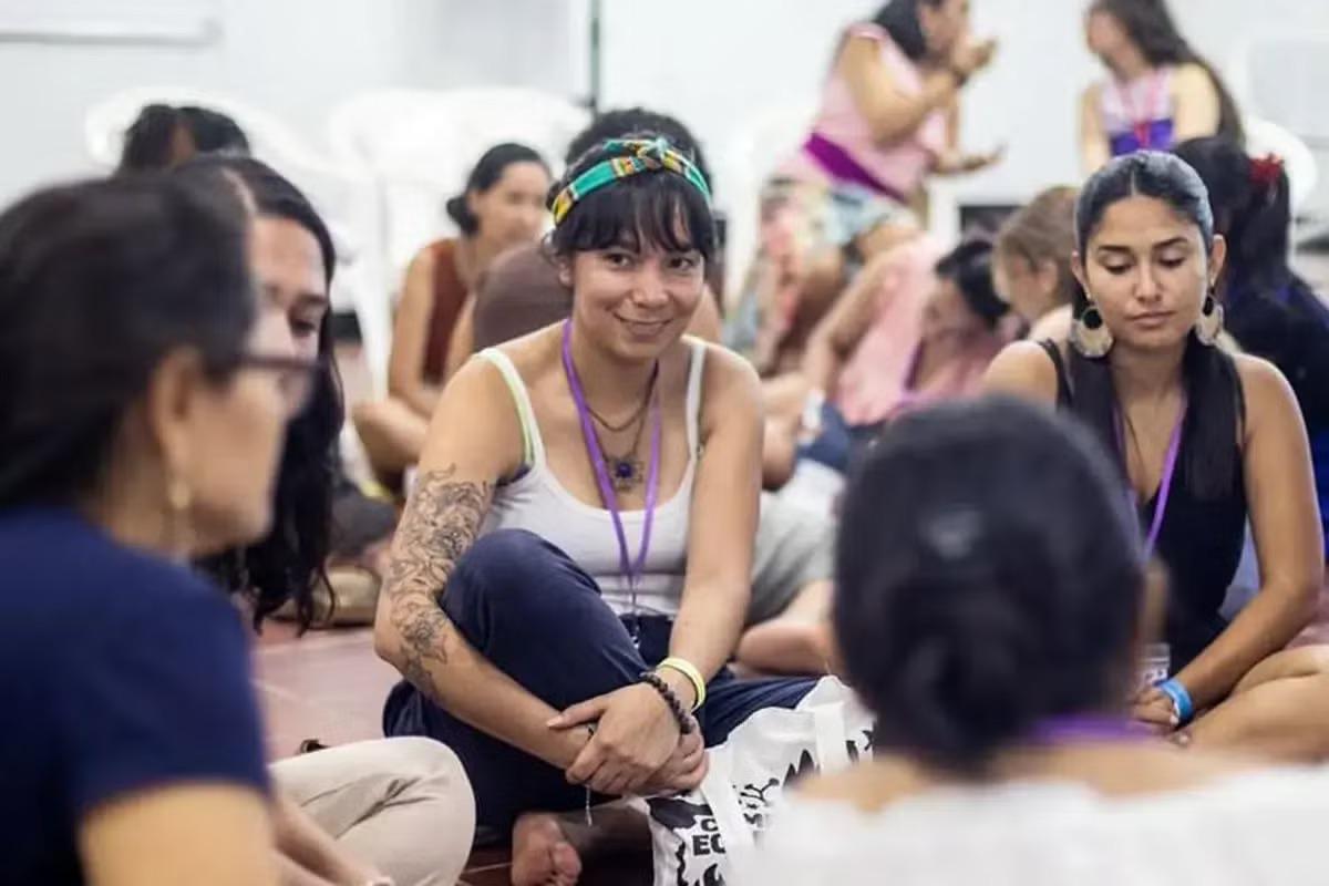 Un grupo de mujeres sentadas, con una joven sonriente en el centro que lleva pañuelo en la cabeza y un tatuaje visible en el brazo.