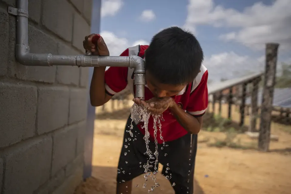A young boy in a red shirt drinks water from a tap attached to a wall.