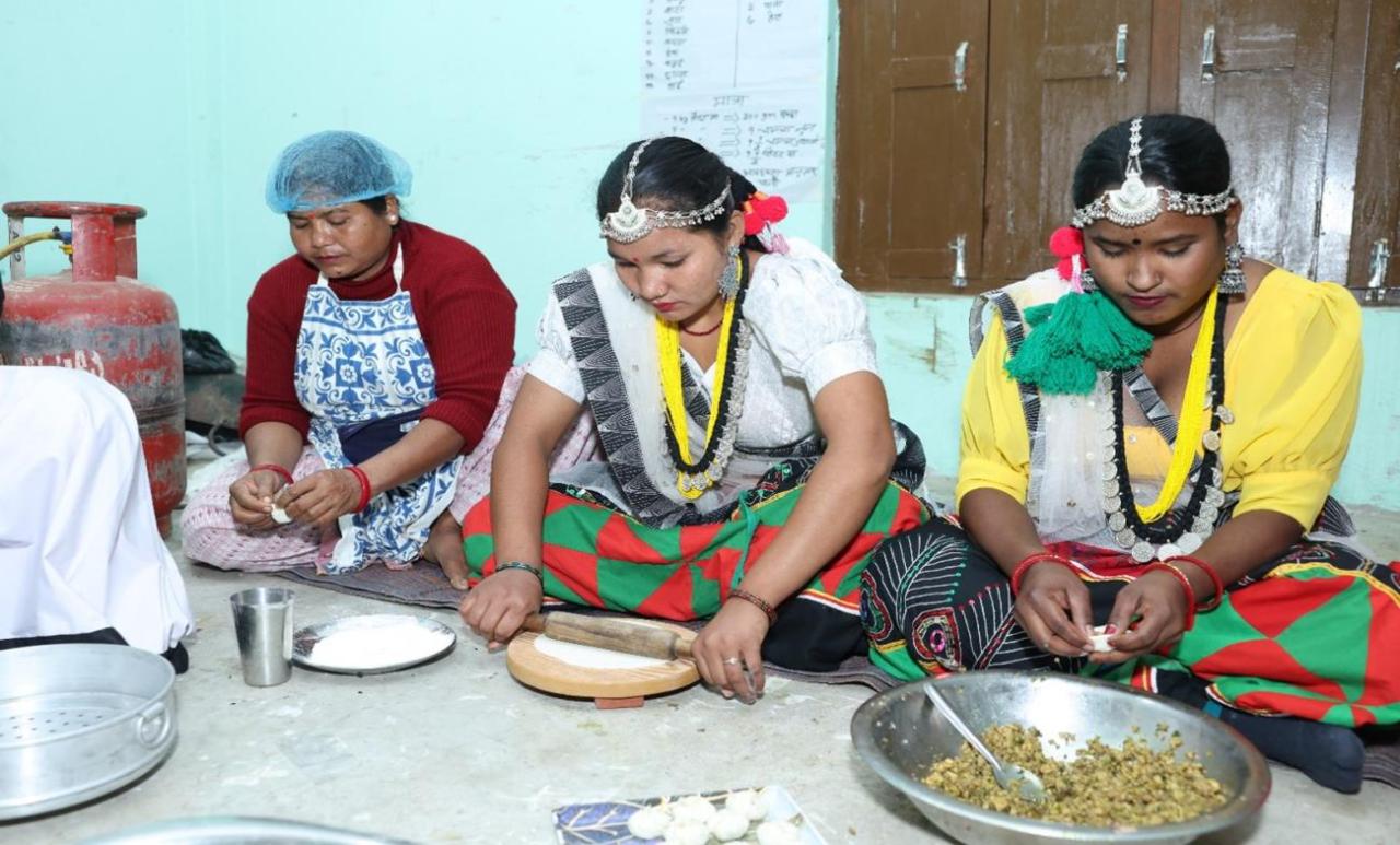 A group of women in traditional clothing eat from plates, while sitting on the floor.