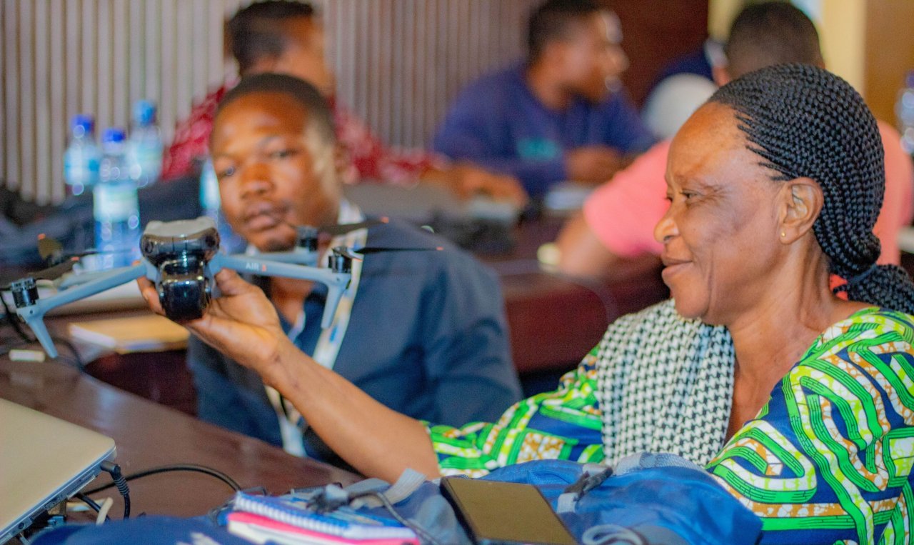 A participant in a workshop holds and examines a small drone while seated at a table with others in the background.
