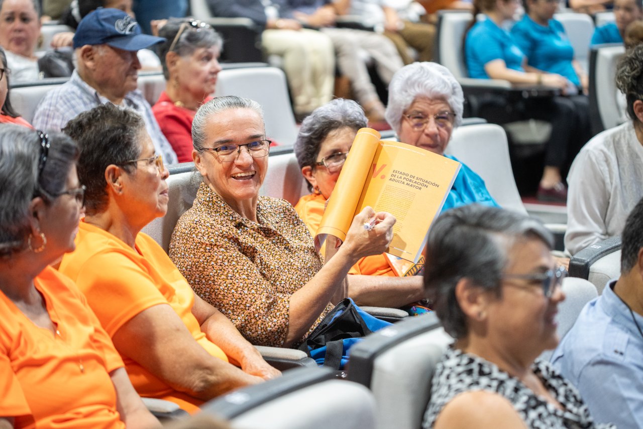 Audience members seated in a lecture hall review a printed report titled “Estado de Situación de la Población Adulta Mayor.”