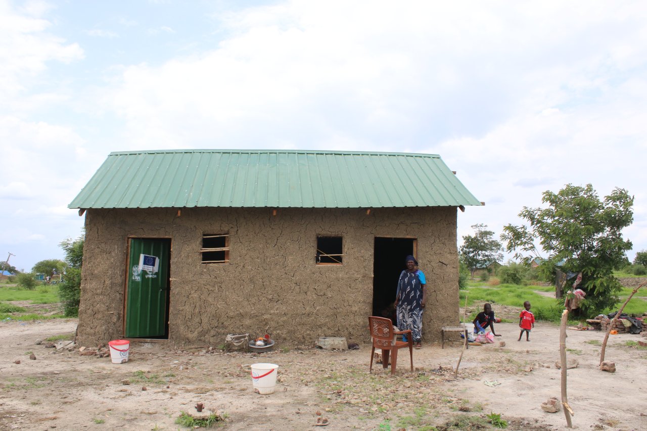 A wide shot of a woman standing in front of a house in South Sudan.
