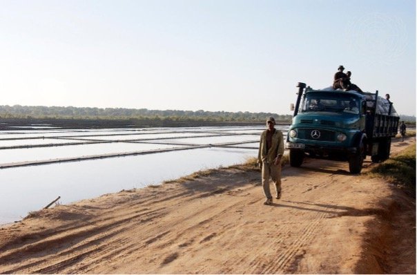 Un hombre camina por un camino de tierra junto a unas salinas, mientras detrás de él avanza un camión con varias personas a bordo.