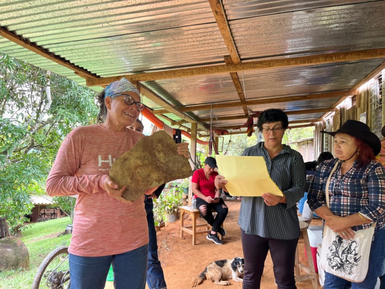 Varias mujeres participan en una actividad comunitaria bajo un cobertizo; una sostiene una gran pieza de material natural mientras otra lee un documento.