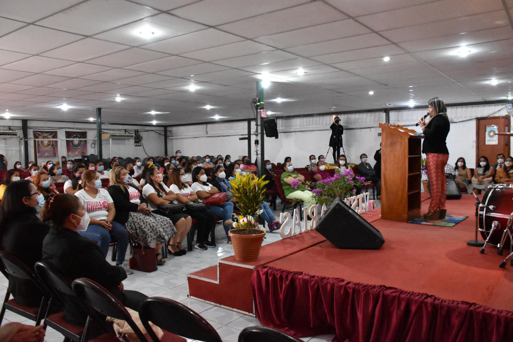 A woman speaks at a podium in front of a large group of women in face masks.