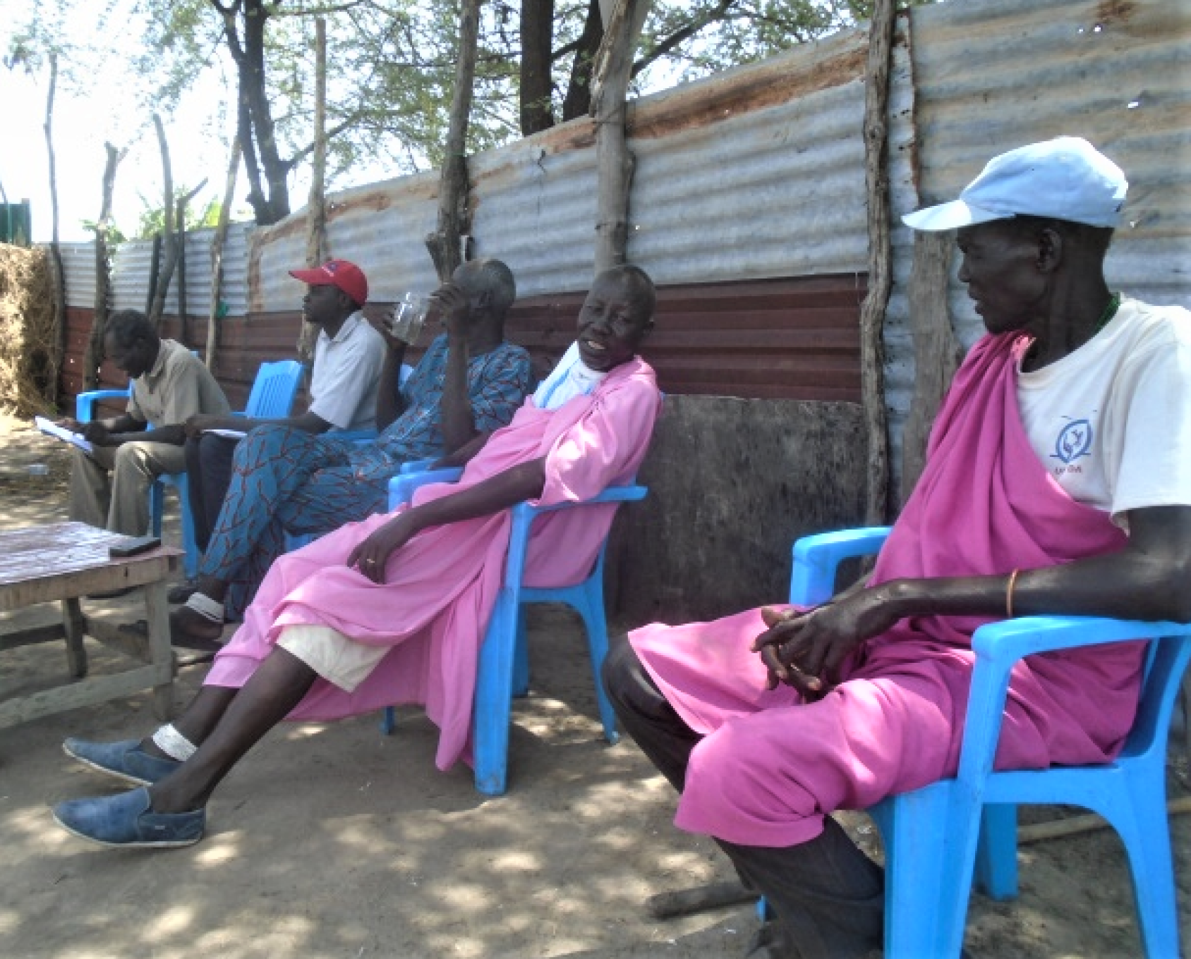 Several people sit in blue chairs near a metal fence. 