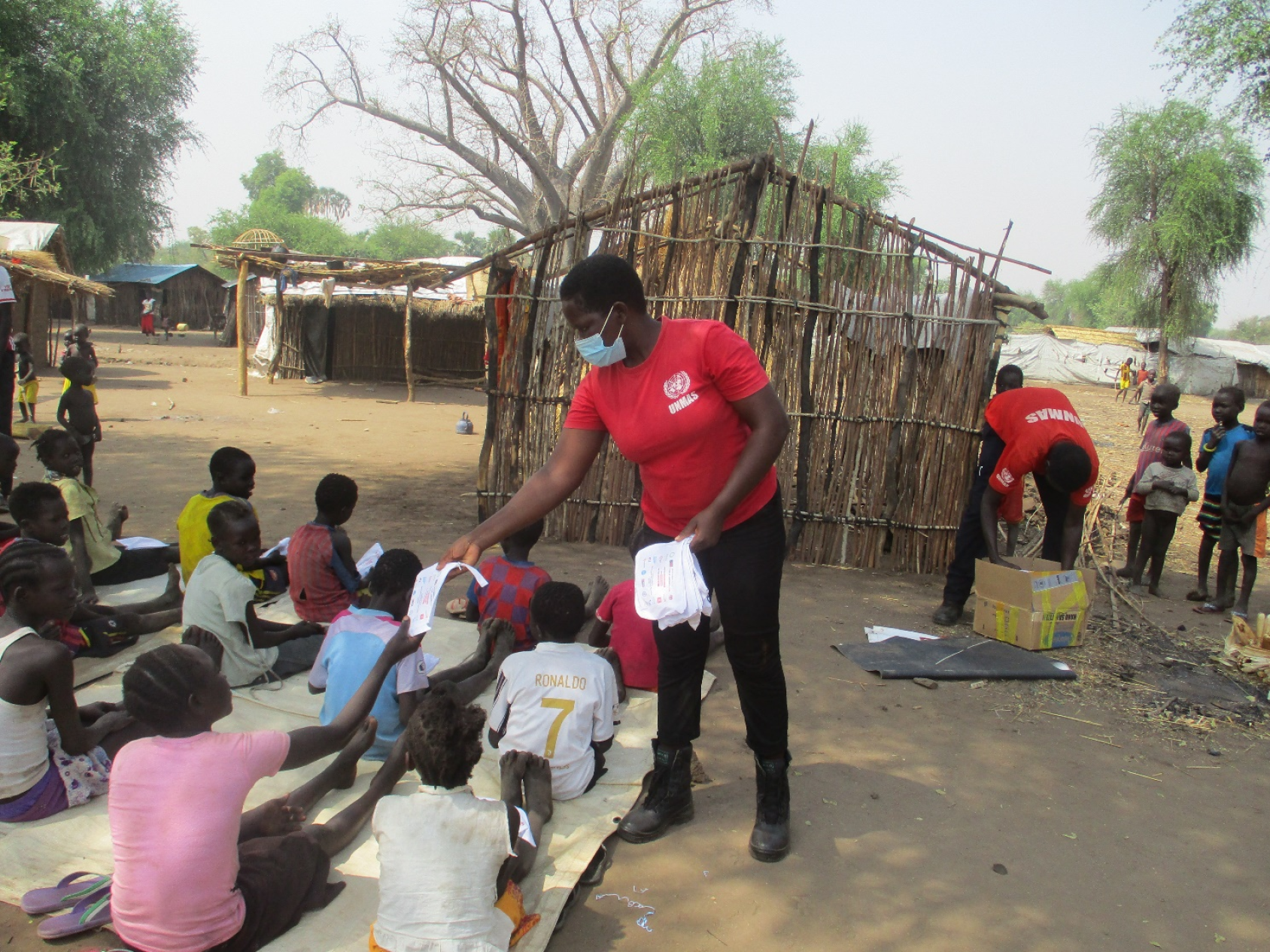 A woman in a red shirt and light blue face mask hands out information to a large group of children seated on the ground. 