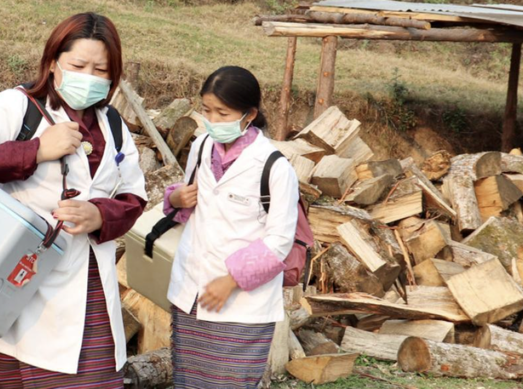 Two women in medical attire and light blue face masks stand near a pile of wood. 