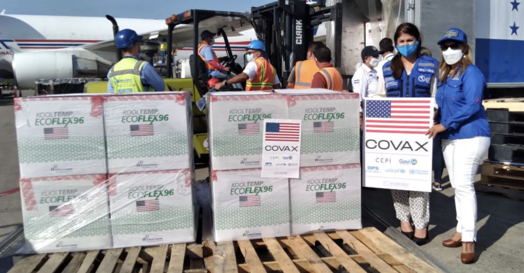 يقوم عدد من الأشخاص بتفريغ لقاحات كوفيد-19 من الطائرات، بينما تحمل امرأتان لافتة تابعة لمرفق كوفاكس.