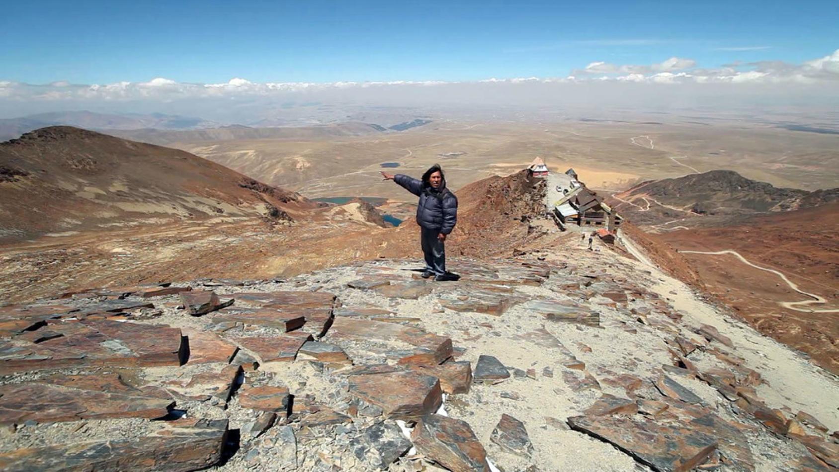 Cumbres de una región montañosa, con una persona usando ropa de invierno y señalando una cumbre del árido paraje natural, el cual en otros tiempos fue una estación de esquí.