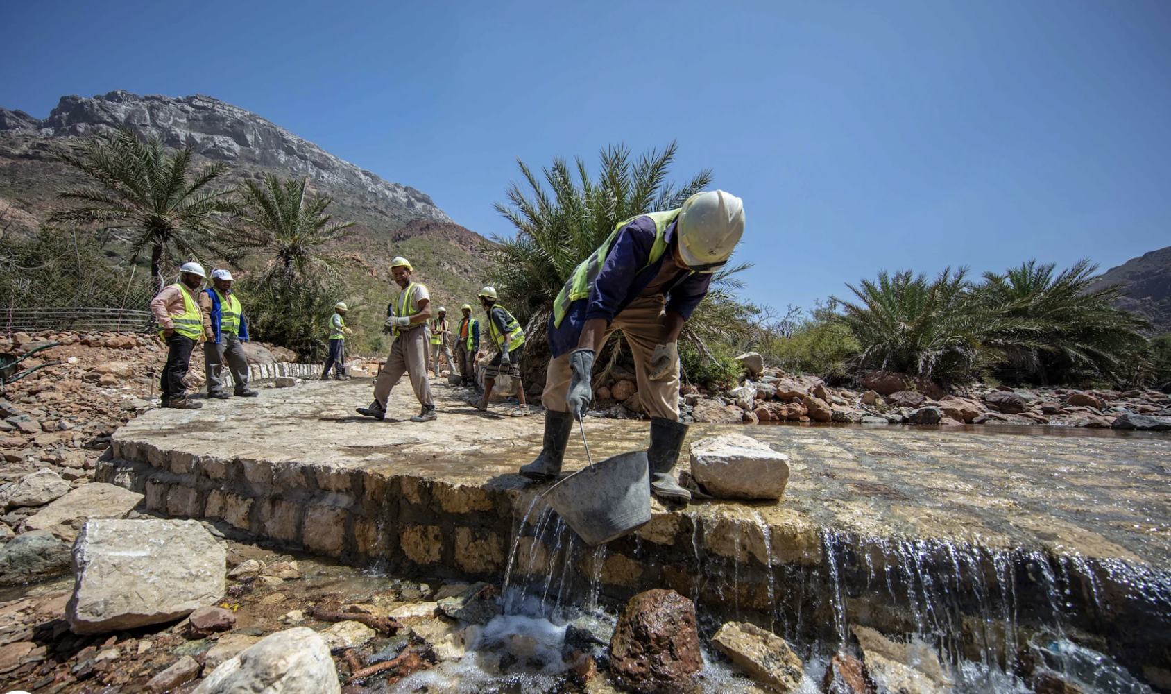 Hombre con un balde de agua, agachado hacia la cámara mientras realiza trabajos de reparación de la carretera.