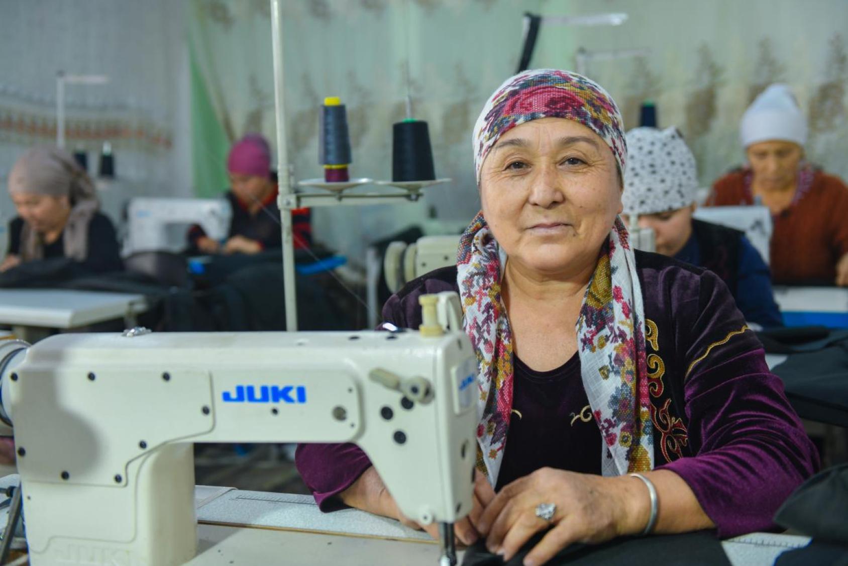 Une femme est assise devant une machine à coudre, dans un un atelier où travaillent plusieurs femmes couturières. Elle regarde l'objectif avec un léger sourire.