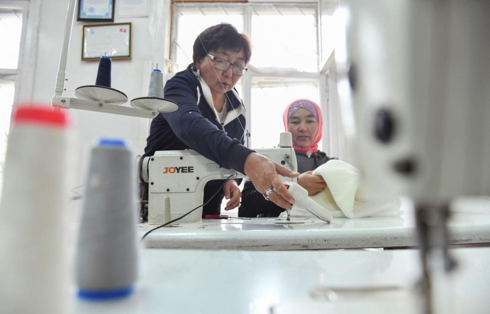 Dans un atelier de couture, une femme aide une autre femme à utiliser une machine à coudre blanche. 