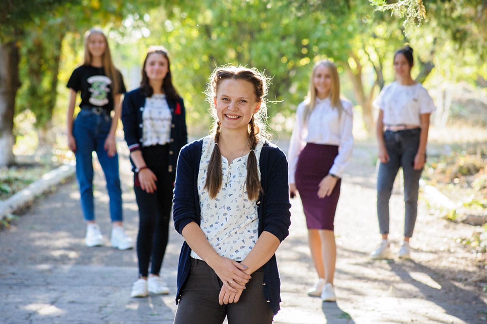 Five girls stand, socially distanced, outside near some green trees. 