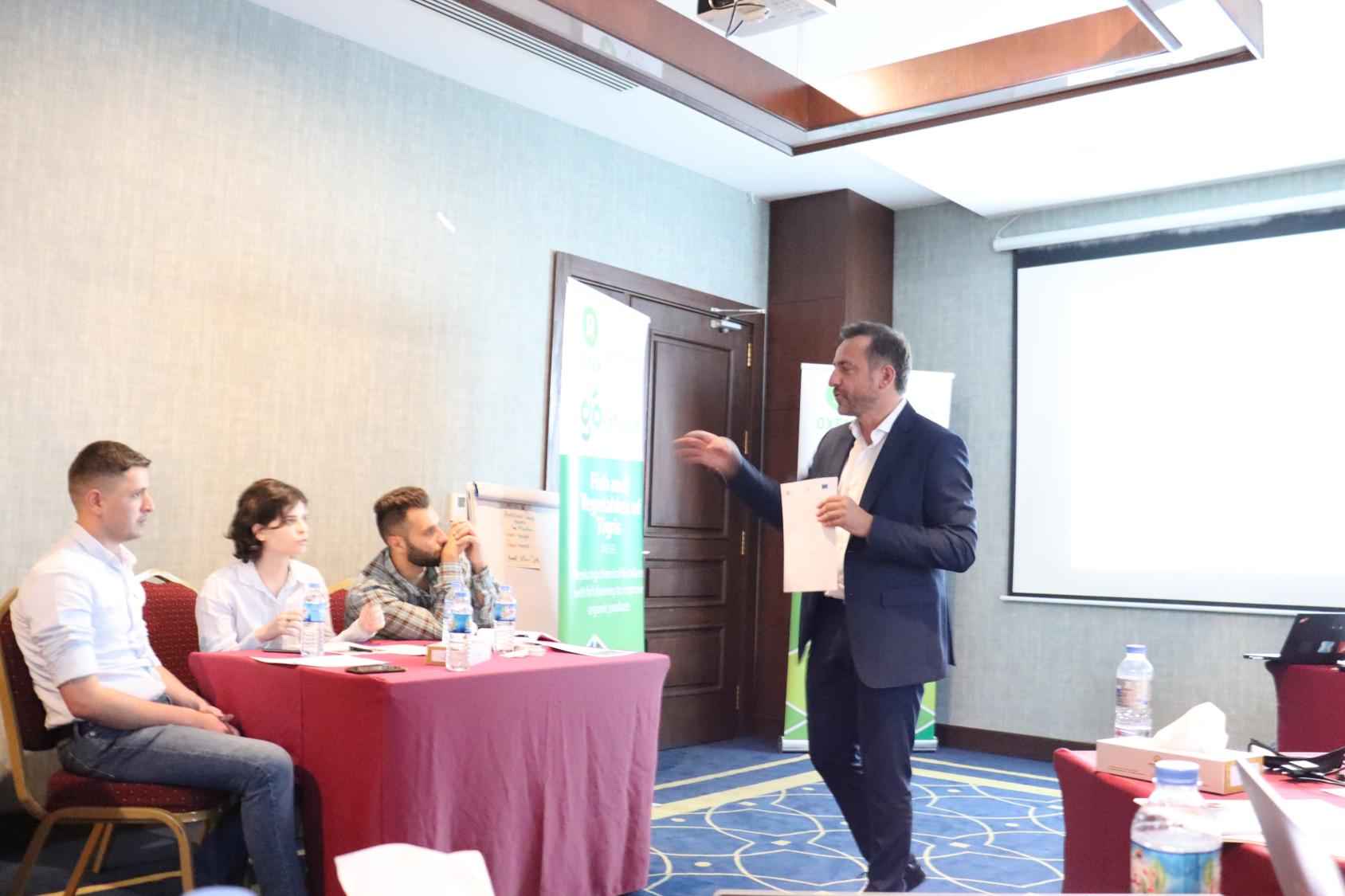 رجل يتحدث إلى ثلاثة أشخاص يجلسون على طاولة إلى يمينه.