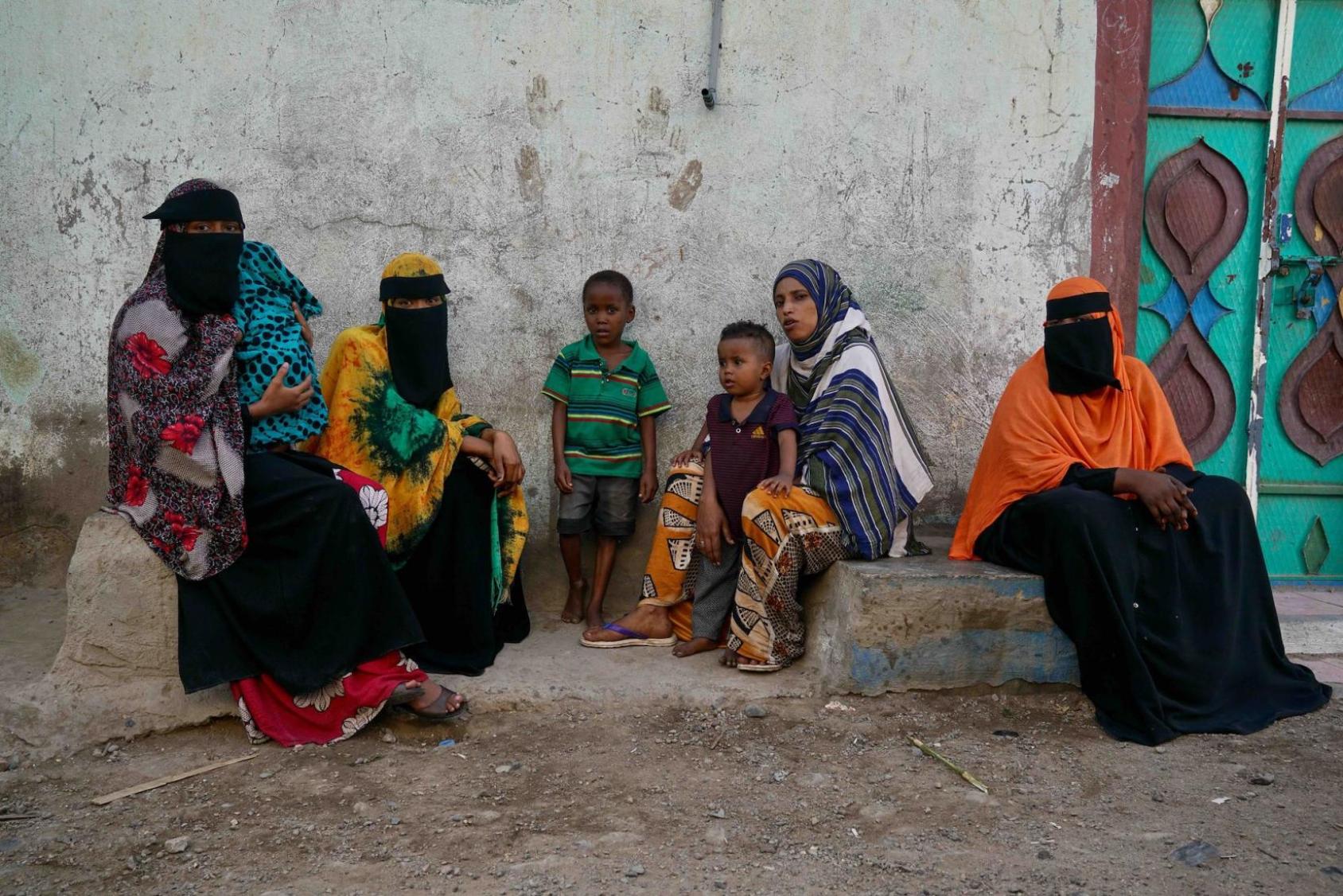 A group of women and children sit on the side of a dirt road. 