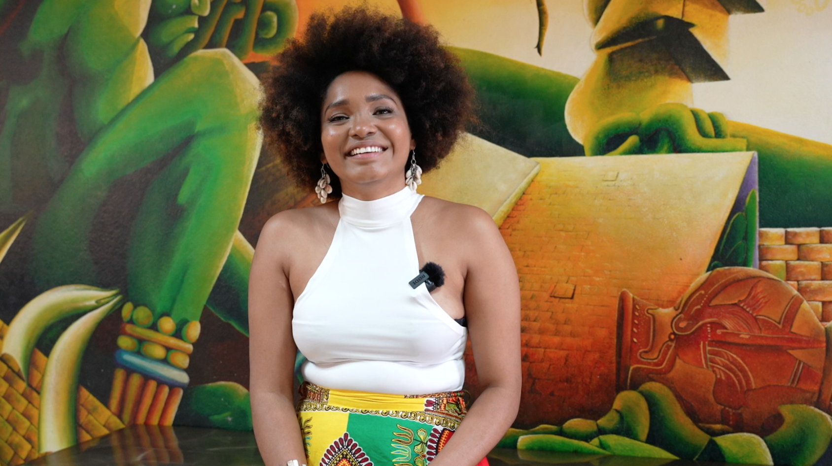 امرأة تبتسم للكاميرا وهي تجلس أمام لوحة جدارية ذات ألوان زاهية.