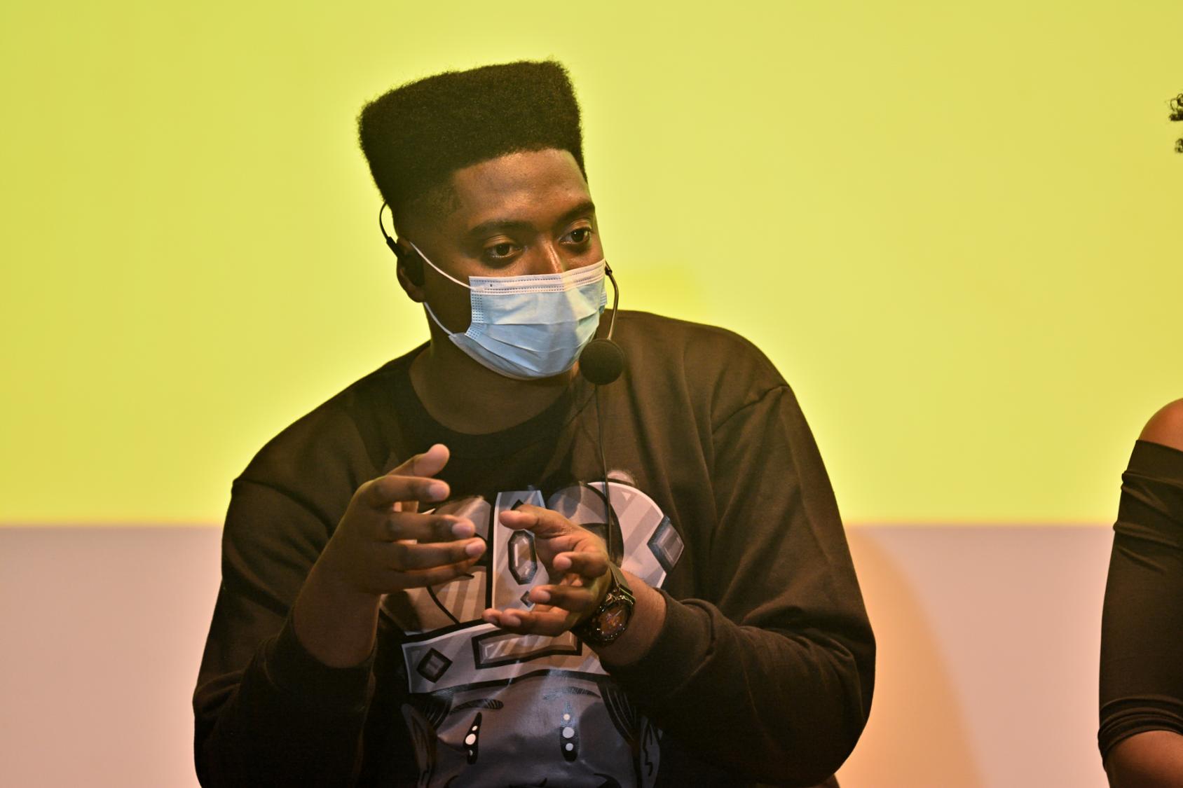 رجل يرتدي كمامة ويتحدث أمام شاشة عرض صفراء.