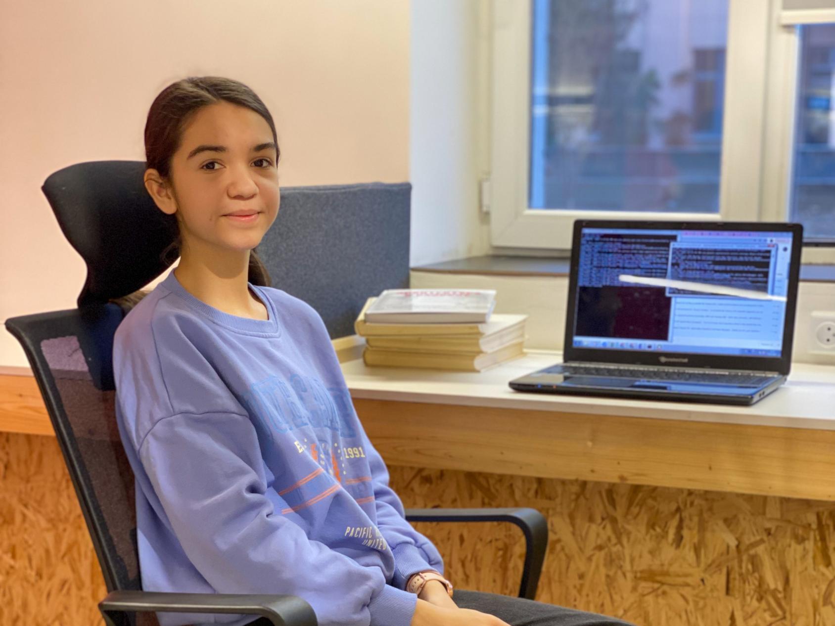 Une jeune fille en pull bleu est assise devant un ordinateur portable à un bureau. 