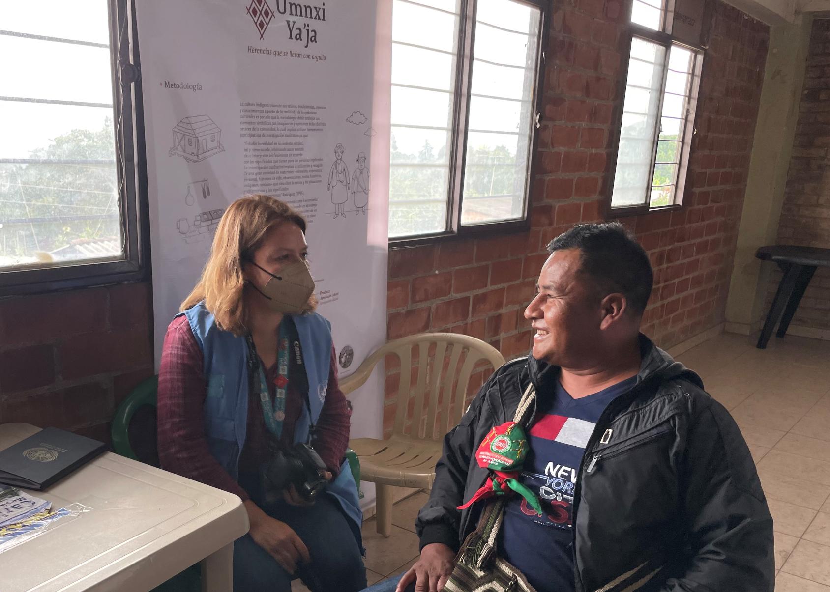 A woman, wearing a blue vest, and a man, in a black jacket, sitting next to each other while they exchange thoughts.