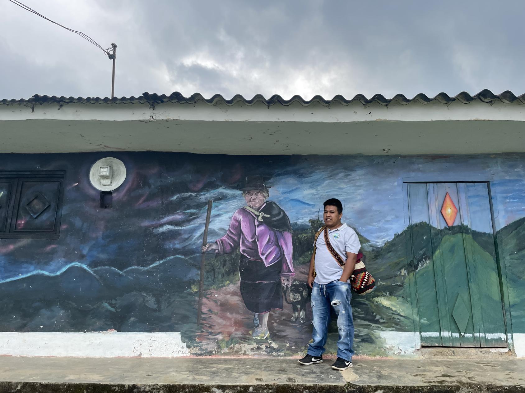 A man is standing in front of a mural that portraits a woman wearing traditional attire in a mountainous landscape.