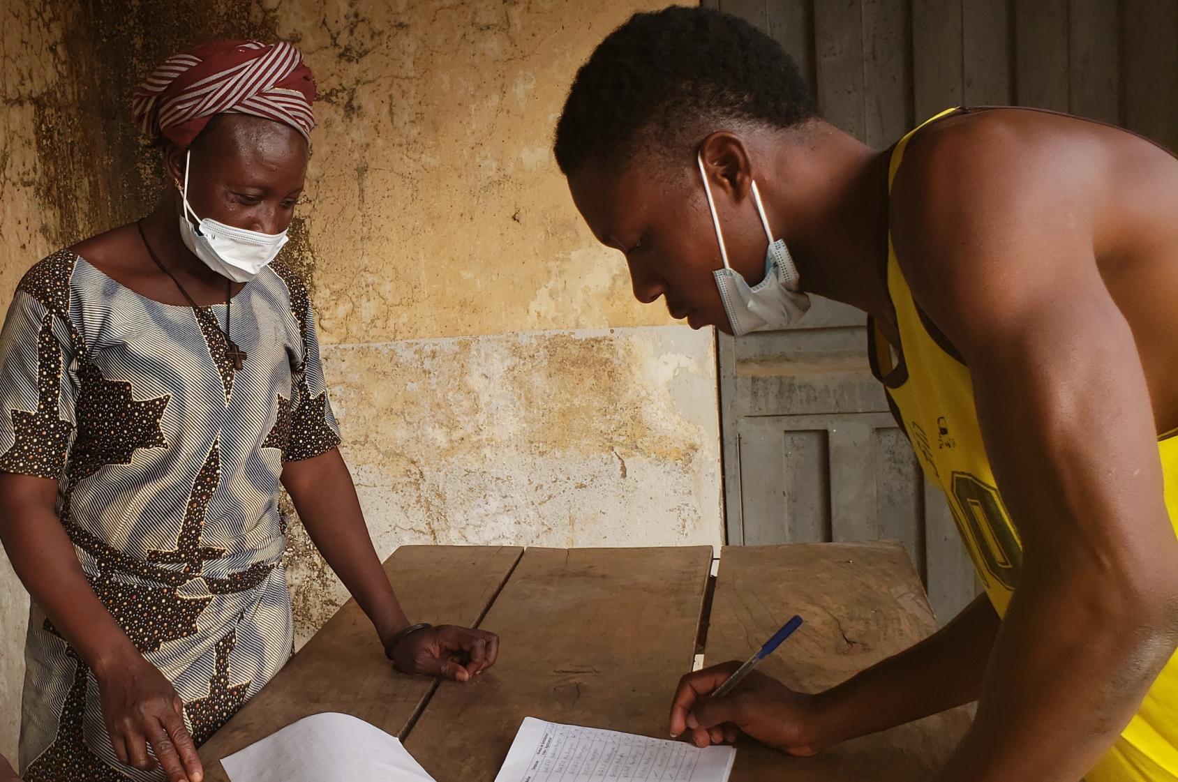 شاب يرتدي قميصًا أصفر بلا أكمام يتكئ على طاولة خشبية ويكتب على ورقة. تقف امرأة على الجانب الآخر من الطاولة.