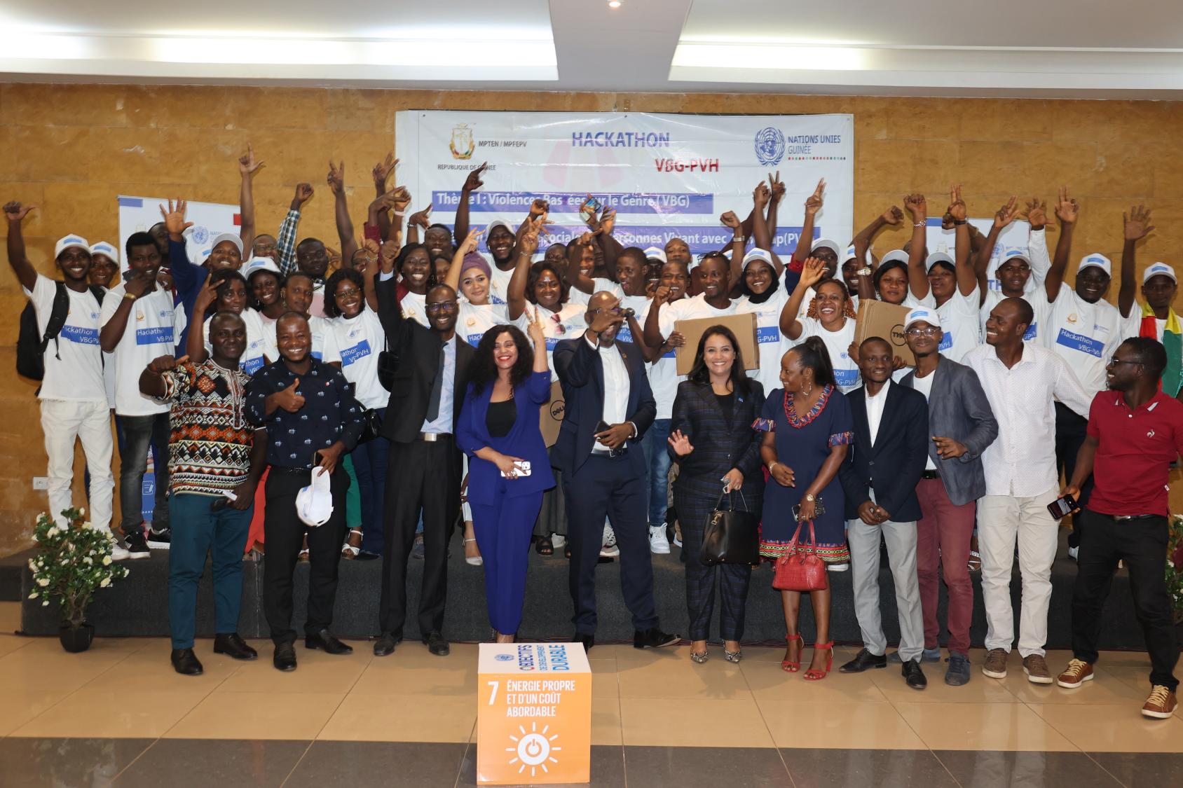 Une trentaine de personnes regroupées sur une estrade posent devant la caméra en levant la main et en souriant.