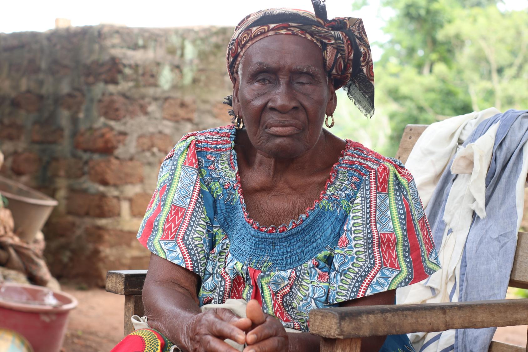 امرأة مسنة ترتدي فستانًا ملونًا وشعرها مربوط من الخلف بوشاح، وتجلس على مقعد خشبي وتنظر إلى الكاميرا.