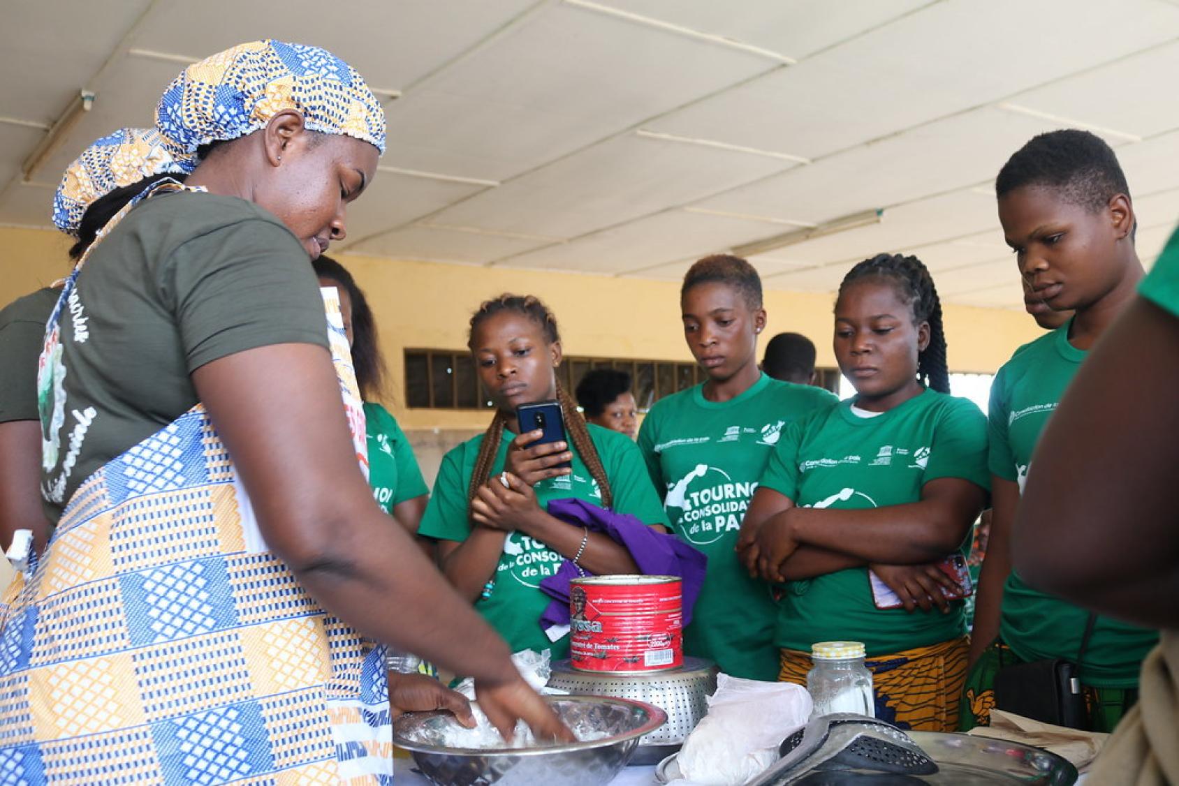 امرأة ترتدي مريلة مطبخ تستخدم الأواني أمام شابات يراقبنها بدقة.