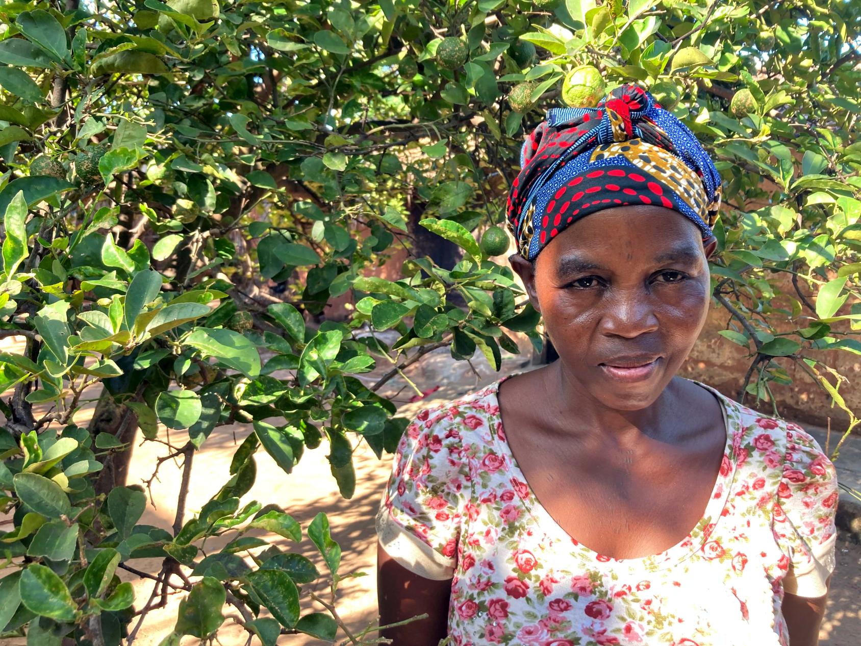 امرأة ترتدي فستانًا منمقًا وغطاء رأس ملون تبتسم وتقف في مواجهة الأشجار الخضراء.